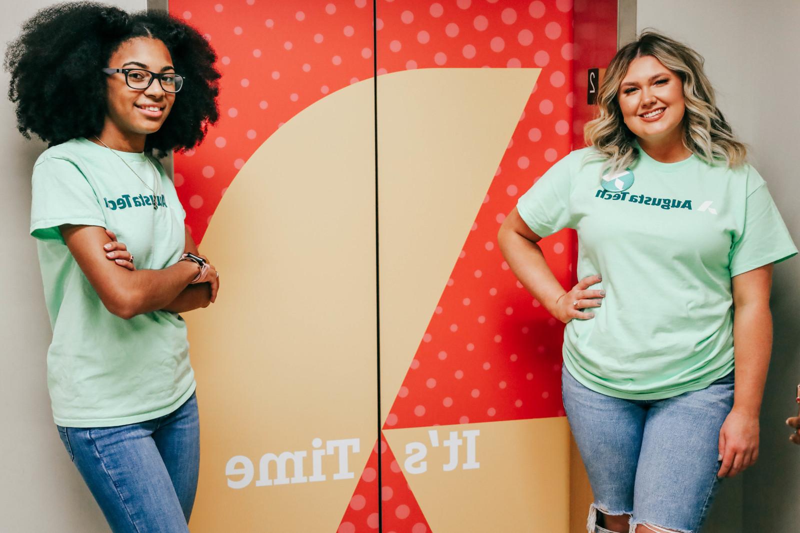 Two female students one a Cacasian female with long blonde hair standing on the left and the other an African American female with shoulder length natural hair on the right, stand wearing mint green 买世界杯app推荐 shirts in front of the 1400 bldg elevator marked by an orange background with a yellow 买世界杯app推荐 A icon and the motto It's Time.