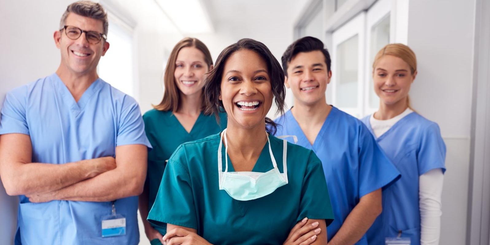 Five health care workers stand in a hallway wearing scrubs in various shades of blue. An African American female stands center with 白人男性 and female standing directly behind her. Another Caucasian female stands to the far left behind the Caucasian male and another tall Caucasian male stands to the far right.